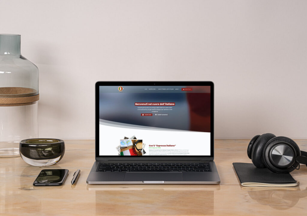Un computer portatile su cui è visualizzato un sito web italiano, probabilmente Sito Espresso, è appoggiato su una scrivania di legno, insieme ad alcune cuffie, un telefono, un quaderno, una penna, una ciotola e un barattolo di vetro decorativo.