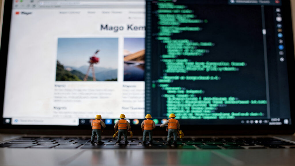 Quattro statuine di operai edili posizionate sulla tastiera di un laptop di fronte allo schermo che mostra codice e un sito web